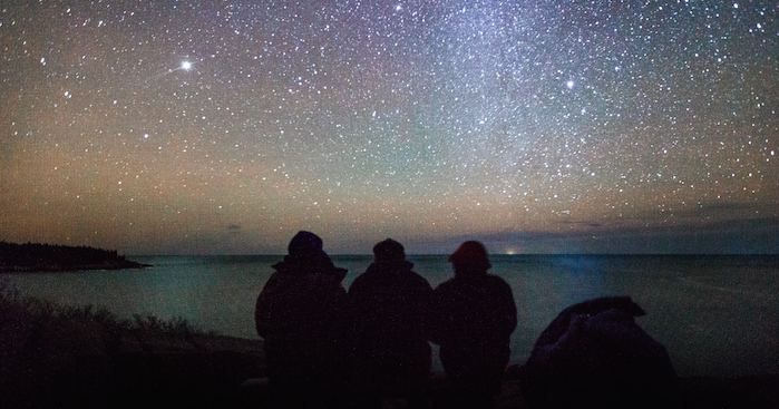Three people sit looking over the water at Acadia National Park. The night sky is filled with star and you can see a hint of he Milky Way.