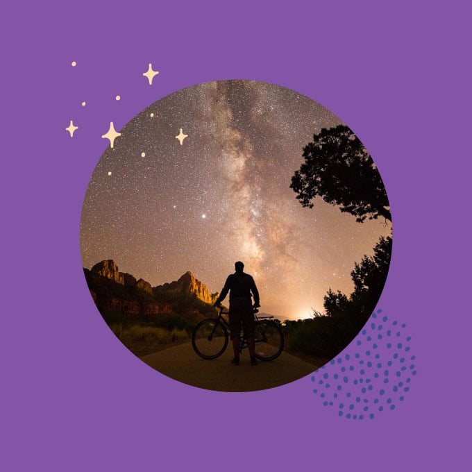 Photo illustration of a silhouetted person standing with their bicycle on a dark path at night, with the Milky Way showing brightly in the background.