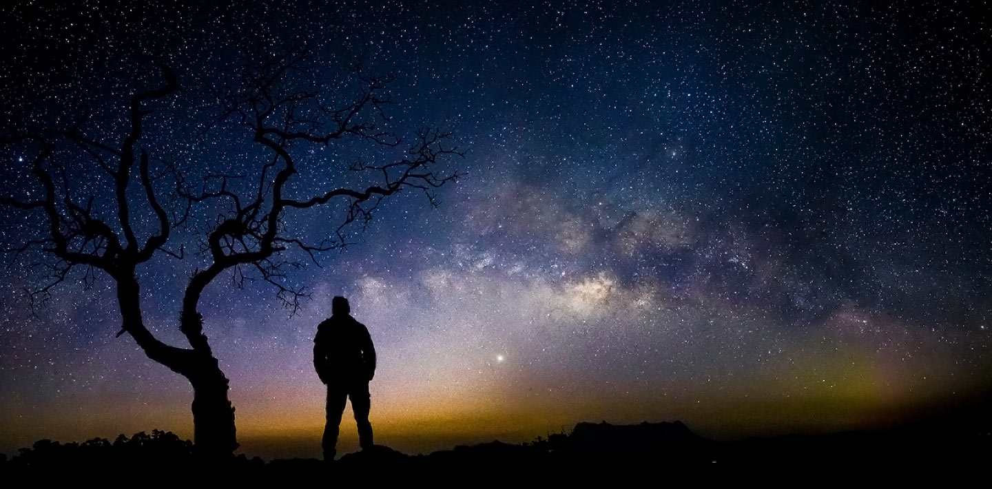 Silhouette of a man and tree against a brilliant, colorful Milky Way night sky.