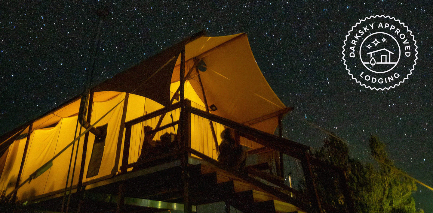A tent cabin at night, glowing slightly with yellow light, while the stars shine above.